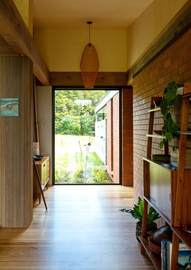 A warm, narrow residential hallway or entry passage with polished timber flooring, exposed timber beams and ceiling, a red brick feature wall, vertical timber wall panelling, and a large black-framed glazed door opening onto a garden. Mid-century-style timber shelving and cabinetry line the right wall, with indoor plants and a sculptural pendant light adding natural character.