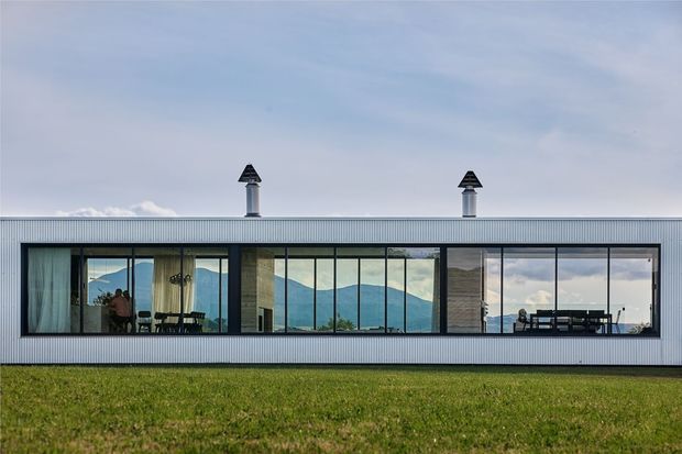 A long, low contemporary building with a corrugated metal facade and a shallow flat roof, viewed from across a grassy lawn. A continuous band of dark-framed floor-to-ceiling glazing reveals interior dining and living areas, with mountain scenery reflected and visible through the windows. Two roof-mounted metal chimneys punctuate the simple horizontal form.