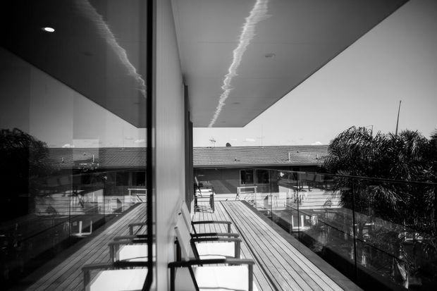 Black-and-white architectural photograph of an elevated timber deck or balcony running beside a glazed building facade. Outdoor chairs line the edge, with clear glass balustrades, a broad projecting roof canopy, and views toward a low tiled-roof structure and palm vegetation. Strong reflections in the glazing emphasize the linear modern composition.