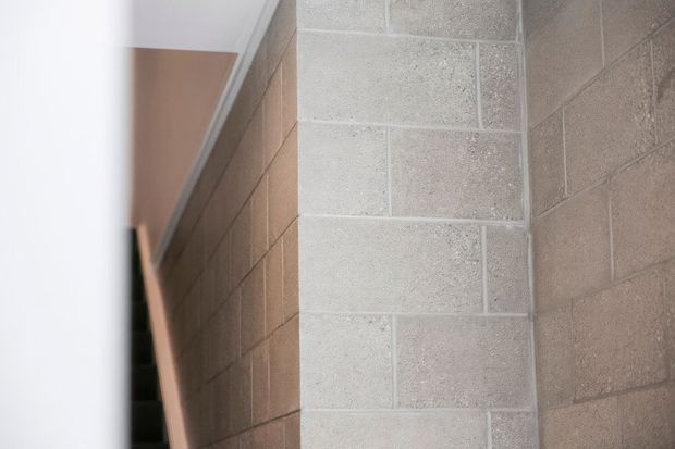 Close-up interior view of a masonry block wall forming a corner beside a narrow stair or passage. The walls are constructed from exposed light grey and taupe concrete masonry blocks with visible mortar joints, and a white ceiling edge is visible above. The composition is minimal and architectural, focused on the textured blockwork and circulation space.