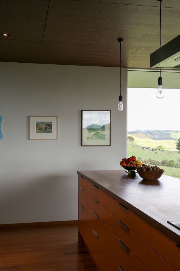 A warm, minimalist kitchen interior with a large central island featuring extensive orange-brown built-in drawer cabinetry and a smooth pale benchtop. The room has a dark timber-lined ceiling, timber flooring, plain light walls, pendant lights, framed artwork, and a large opening overlooking rolling rural farmland. The composition emphasizes the island, natural light, and landscape connection.