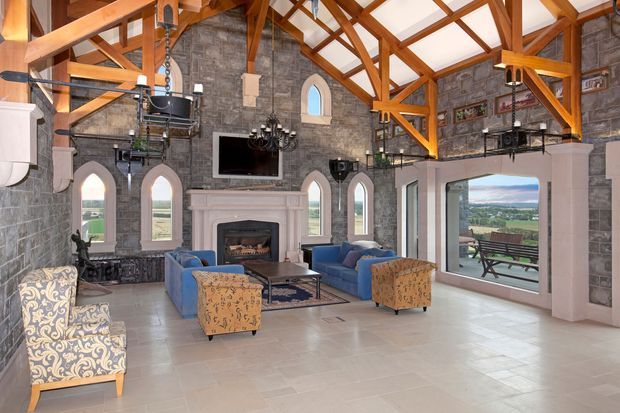 A spacious double-height living room with a dramatic vaulted timber-trussed ceiling, exposed structural beams, and grey stone masonry walls. Tall arched windows and doors bring in expansive rural views. The room includes a stone fireplace with TV above, chandeliers, pendant lights, blue sofas, patterned armchairs and ottomans, and a light tiled floor. The overall character blends rustic stone architecture with refined lodge-style furnishings.