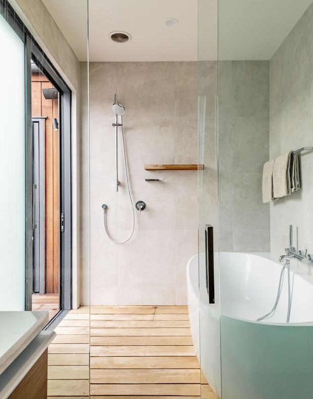Bright contemporary bathroom with a walk-in glass shower, pale large-format wall tiles, timber slatted flooring, and a freestanding white bathtub with chrome fittings. A narrow timber shelf and wall-mounted towel rail provide simple functional detailing, while a glazed opening brings in natural light.