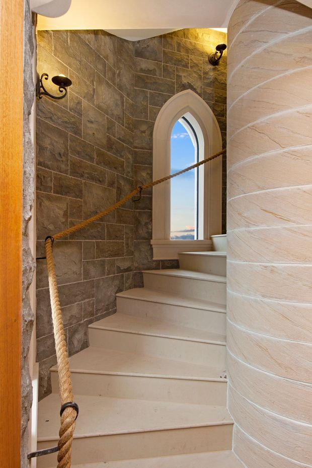 A narrow curved stairway rises beside a cylindrical textured plaster or stone column. The walls are clad in irregular dark grey-brown stone tiles, with warm wrought-iron wall sconces and a tall pointed-arch window. A thick rope handrail follows the staircase, creating a distinctive rustic nautical detail. The stairs and architectural trim are painted in a light cream tone, with blue sky visible through the window.