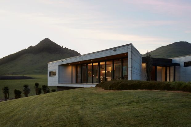A low-slung contemporary single-storey residence set into a broad landscaped hillside, with a crisp rectangular form, flat roof, extensive floor-to-ceiling glazing, and silver-grey horizontal metal cladding. Warm interior lighting glows through the glazed living spaces at dusk, while dramatic green mountains form the immediate backdrop.