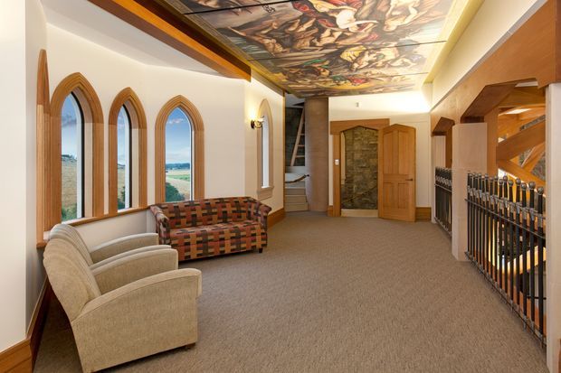 A spacious, traditionally styled interior hallway or lounge area with a carpeted floor, arched timber-framed windows, and a richly illustrated decorative ceiling mural. Upholstered armchairs and a patterned sofa sit along the window wall, while timber joinery, a stone accent wall, stair access, and decorative metal balustrading define the circulation space. The room has warm wood detailing, cream walls, and views of open landscape through the windows.