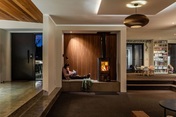 Warm, refined open-plan residential interior featuring a dark entry door, polished concrete entry flooring, a raised carpeted living area, timber ceiling detailing, and a built-in timber-lined fireplace nook with a wood-burning stove. A dining area and extensive built-in bookshelves are visible beyond the living space.