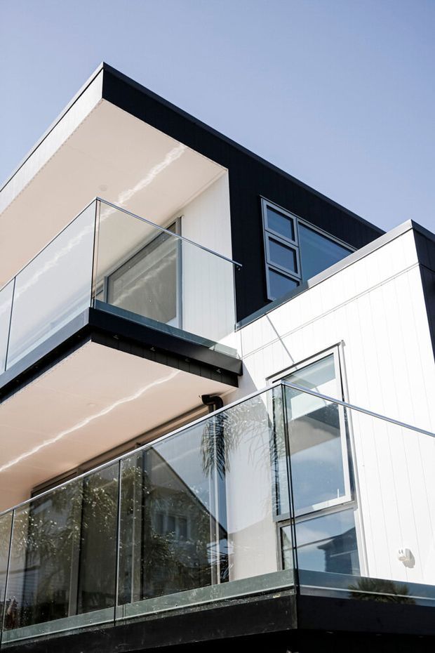 Close-up exterior view of a contemporary multi-storey residential building with strong geometric forms, projecting balconies, frameless glass balustrades, black cladding, white vertical wall panels, and large windows. The image is photographed looking upward against a clear sky.