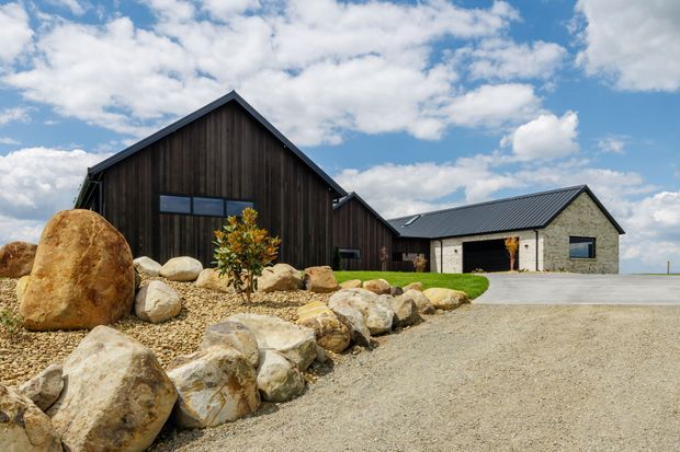 Exterior view of a contemporary rural-style property composed of dark vertical timber-clad gabled volumes connected to a lighter masonry-clad wing. The building sits behind a broad gravel driveway with large natural boulders and sparse landscaped planting. Black metal pitched roofs, expansive dark-framed glazing, and a simple low-profile composition are visible beneath a partly cloudy sky.