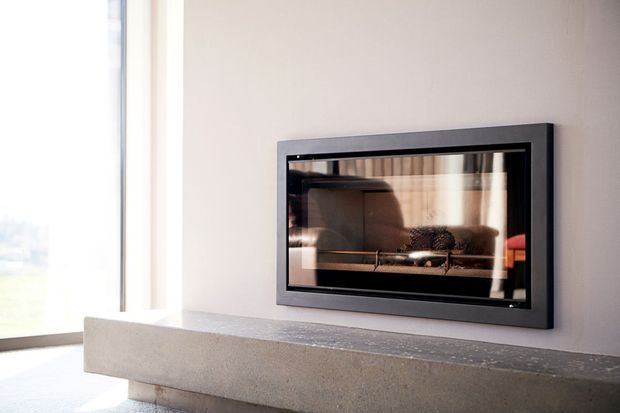 Close-up of a contemporary built-in fireplace with a black metal frame and glass front, set into a light neutral wall above a substantial grey concrete hearth or mantel. Bright floor-to-ceiling glazing is visible at the left, and the fireplace glass reflects the room and curtains.