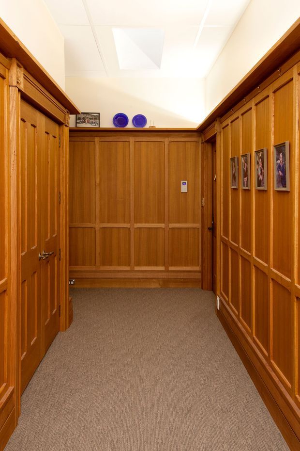 A narrow interior hallway lined with extensive warm-toned timber wall panelling and built-in cabinetry or doors. The corridor has a carpeted floor, framed artwork, a small bench-like ledge at the far end, and a white suspended ceiling with a rectangular skylight or light panel. The space has a traditional, richly crafted character.