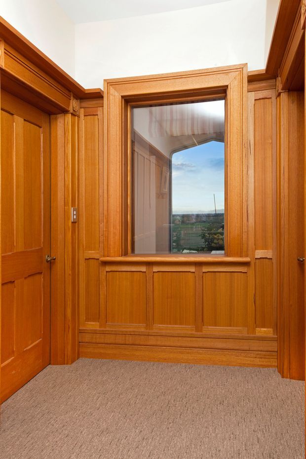 A compact traditional wood-panelled interior hallway or landing with extensive warm honey-toned timber wall panelling, decorative moulded trim, timber doors, and a framed window. The window reflects an exterior view and opens toward a distant landscape. The floor is covered in neutral beige carpet, and the space is brightly lit with a white ceiling.