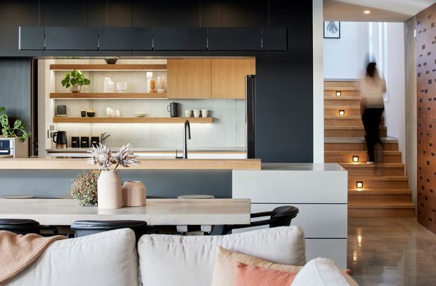 A contemporary open-plan kitchen and dining/living area with dark cabinetry, pale timber shelving and cabinetry, a light stone island bench, and integrated open shelving. The foreground includes a dining table and sofa seating, while timber stairs with illuminated treads lead upward beside a perforated brick feature wall.