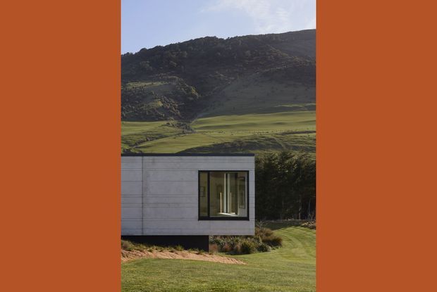 A compact contemporary pavilion or small dwelling set within a manicured rural landscape, with a dramatic dark rocky hillside rising behind it. The visible building has a simple rectangular form, flat roof, pale horizontal-panel exterior cladding, and a large dark-framed window. The image is vertically framed with broad terracotta side borders, emphasising the contrast between the minimalist structure and the green mountainous setting.
