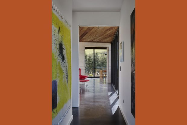 A bright, narrow residential hallway leading toward a dining or living area with floor-to-ceiling glazing. The corridor has smooth white walls, dark polished concrete flooring, a warm timber-lined ceiling feature, dark built-in cabinetry or door panels, and a vivid red chair visible near the glazed end. Artwork adds a bold yellow and black accent along the left wall.
