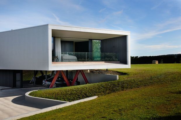 Modern cantilevered rectangular house set in an open grassy landscape. The upper volume has pale ribbed cladding, a deep recessed glazed balcony or living area, and dark vertical wall cladding. A dramatic red structural support frame lifts the upper level above a partially enclosed garage or lower level, while curved concrete paving and landscaped lawn surround the building.