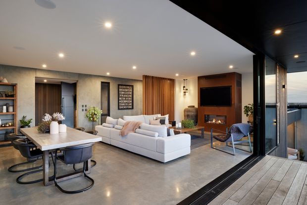 Open-plan contemporary living and dining area with polished concrete flooring, a large stone dining table, white sectional sofa, timber feature wall, built-in fireplace and television joinery. Full-height black-framed glazing opens onto a timber deck, creating a bright indoor-outdoor connection.