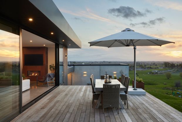Contemporary rooftop-style terrace with a timber deck, outdoor dining table and chairs, and a large cantilevered umbrella. Full-height glazing opens to a warm modern living room with a fireplace, while the elevated terrace overlooks a broad green rural landscape at sunset.