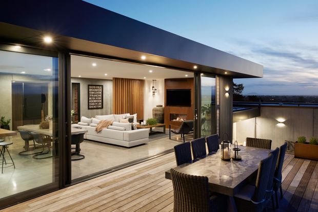 Contemporary rooftop terrace overlooking a distant urban landscape, with a large outdoor dining table and chairs in the foreground. Expansive sliding glass doors open into a refined living room featuring a white sectional sofa, timber slat feature wall, built-in television joinery and warm recessed lighting. The home has a broad flat roof overhang, dark metal framing and timber decking, creating a seamless indoor-outdoor entertaining area at dusk.