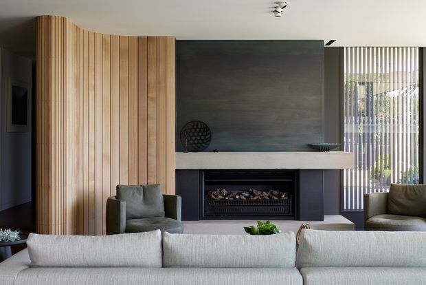 A contemporary living room centred on a built-in fireplace and dark feature wall. A curved vertical timber screen divides the room, while a pale sectional sofa and armchairs face the fireplace. Vertical blinds, restrained decor and a muted neutral palette create a refined, calm interior.