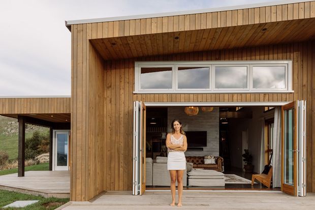 Modern single-storey residential building with a warm vertical timber-clad facade and deep recessed entry terrace. Large bi-fold glazed doors open from the exterior deck into a contemporary living room, with high horizontal clerestory windows above. The simple boxy form, projecting roofline, timber soffit, and connection to the landscaped site are clearly visible.