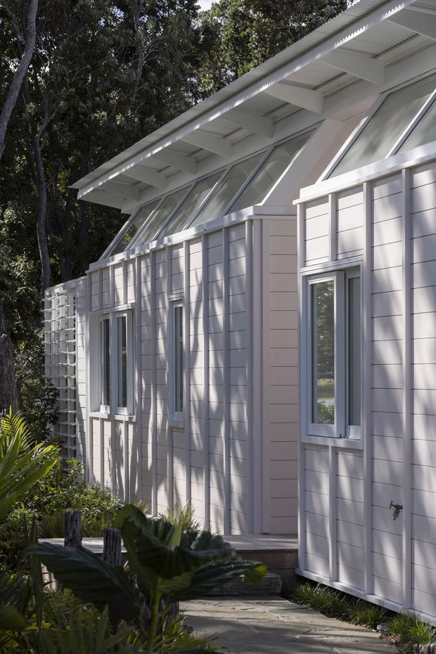 Exterior view of a light-coloured, single-storey building set among mature trees and landscaping. The facade features pale horizontal weatherboard cladding, white-framed windows, a projecting roof with exposed structural rafters, and a distinctive row of sloping clerestory-style windows beneath the eaves. A paved walkway leads alongside the building.