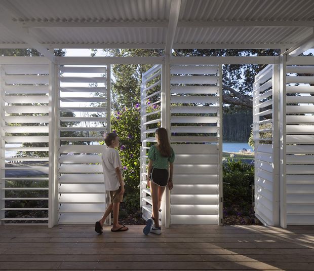 A bright, open-sided outdoor architectural walkway or terrace with a corrugated white roof and multiple white horizontal louvred screens forming partitions and openings. Two people stand on a timber deck, with landscaped vegetation and a glimpse of blue water beyond. The design emphasizes shade, ventilation, privacy, and framed outdoor views.