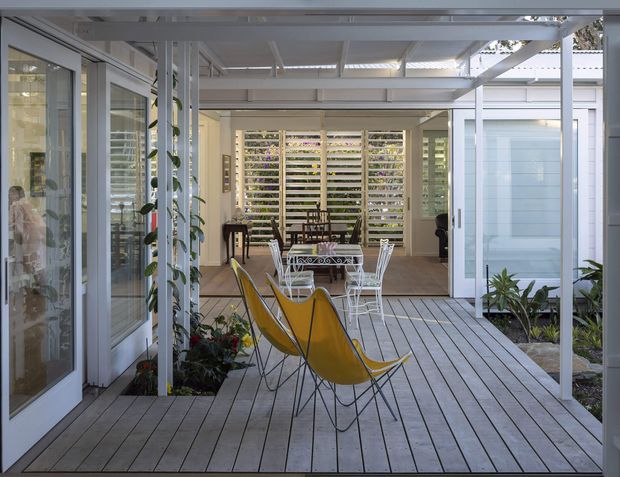 A bright, enclosed outdoor living area with pale timber decking, white-framed glazing, a lightweight roof structure, and louvred screening. The space includes yellow sling lounge chairs, a small white outdoor dining set, potted plants, and a view into an adjoining dining or living area. The design has a relaxed contemporary indoor-outdoor character.