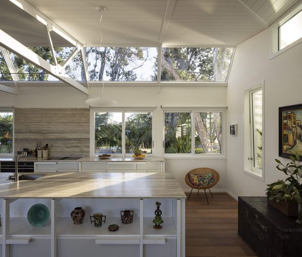 Bright open-plan kitchen interior with a large pale stone island, white open shelving, integrated cabinetry, and a textured stone backsplash. Extensive windows and clerestory glazing overlook leafy trees, while a pitched white-lined ceiling and exposed angled structural elements create an airy architectural character. Timber flooring, sculptural pendant lighting, and a colourful woven accent chair add warmth and detail.