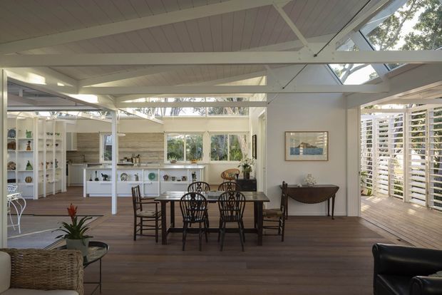 Bright, open-plan dining and living space with a high pitched white timber-lined ceiling, exposed white beams, extensive glazing, and louvred screens opening toward a timber deck. A dark wood dining table with classic spindle-back chairs anchors the room, while built-in white shelving and a kitchen are visible beyond. The interior combines warm timber flooring, white architectural surfaces, and abundant natural light.