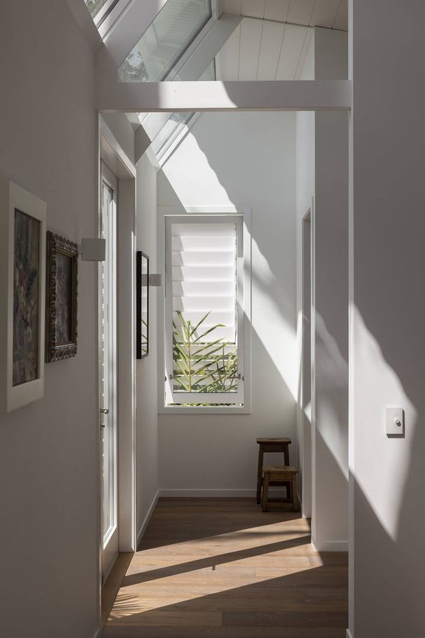 A bright, narrow residential hallway with a tall glazed door, clerestory glazing, and a high ceiling that brings in strong natural light. White walls and ceiling are contrasted by warm timber flooring. A louvred window, framed artwork, wall lights, and a small wooden stool line the corridor, creating a calm, refined interior with pronounced geometric shadows.