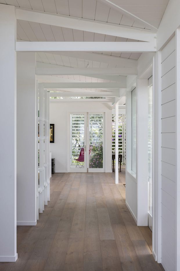 Bright, airy interior hallway with white painted shiplap walls, exposed white timber beams, and a white paneled ceiling. Wide light timber floorboards lead toward glazed doors and louvred windows opening to greenery, creating a long, well-lit circulation space with a coastal character.