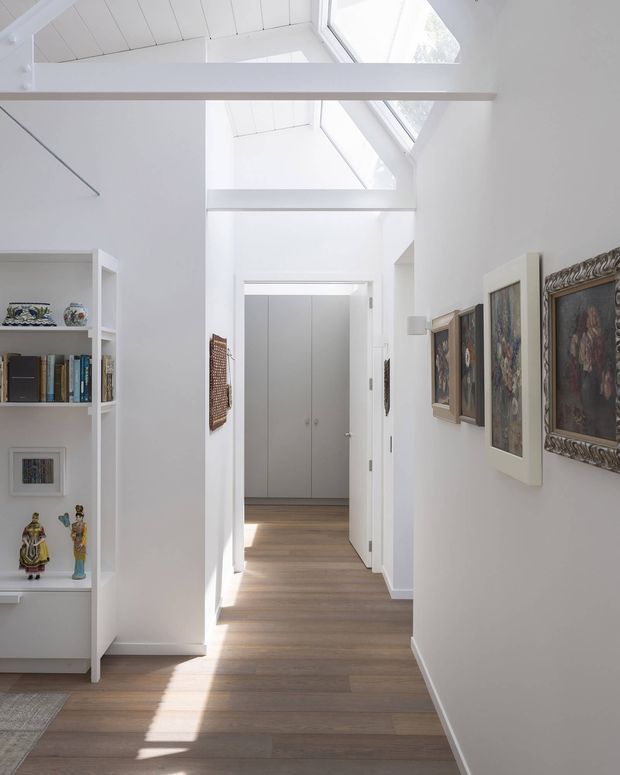 Bright, minimalist residential hallway with white walls, built-in open shelving displaying books and decorative objects, framed artwork along the right wall, and light timber flooring. The corridor leads to a concealed storage area and is illuminated by high clerestory-style glazing and roof windows, with exposed white-painted ceiling beams creating a tall, airy volume.