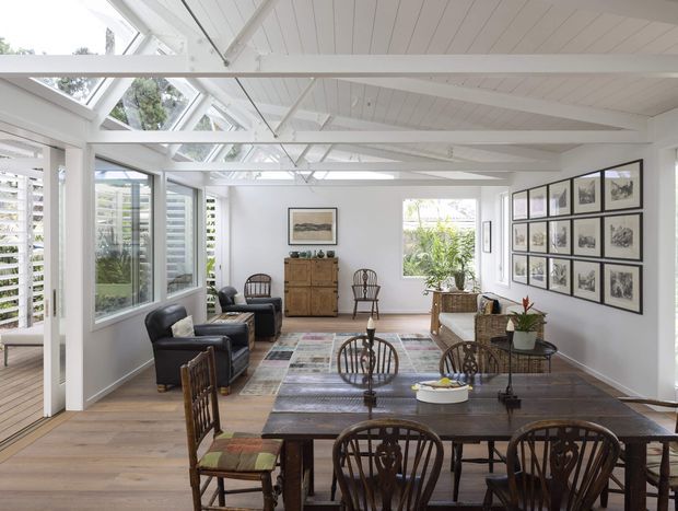 Bright, airy open-plan sunroom or living-dining space with extensive glazing, white-painted exposed roof framing and a pitched glazed roof section. A large dark timber dining table occupies the foreground, with vintage timber dining chairs, leather lounge chairs, a patterned area rug, a timber sideboard, framed artwork, and indoor plants. The room has warm timber flooring and abundant natural light.