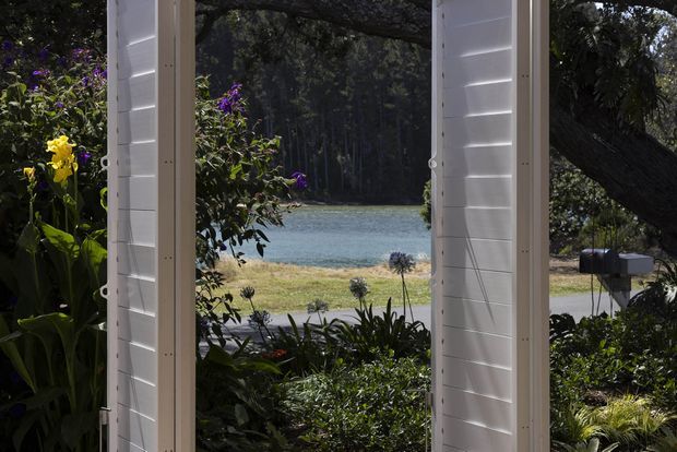 A view through tall white louvred or shutter-style openings toward a landscaped garden, a road, and a calm body of water beyond. Dense tropical and subtropical planting frames the opening, with a large tree canopy and wooded shoreline in the background. The image is predominantly an architectural opening and garden outlook, with strong natural light and contrasting deep shadows.