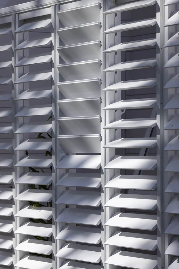 Close-up view of a large modular louvred screen or shading facade composed of repeated white or light-grey horizontal angled slats set within vertical frames. Strong sunlight creates alternating highlights and shadows, with a few small green plants visible behind the screen. The image focuses on the architectural facade system rather than an interior room.