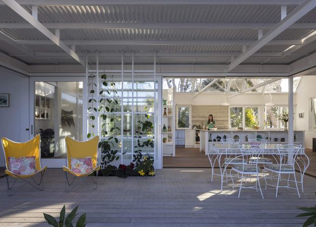 Bright open-plan indoor-outdoor entertaining space with a covered timber deck, white-framed glazing, a dining area, and a kitchen visible beyond. The space features a white exposed beam and corrugated ceiling structure, a glazed or screened garden partition with climbing plants, colourful butterfly chairs, and white metal outdoor-style dining furniture. Abundant glazing and planting create an airy, relaxed atmosphere.