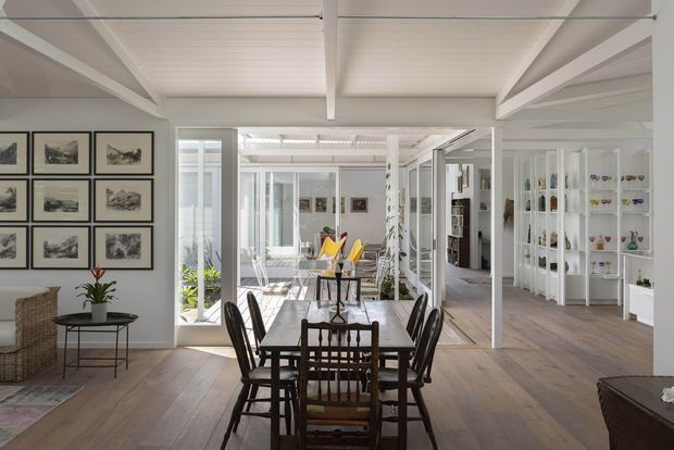 Bright open-plan dining and living space with a timber dining table and dark wood chairs in the foreground, opening into a sunlit sitting area. White-painted exposed ceiling beams and wall framing, large glazed doors and windows, pale timber flooring, built-in white shelving displaying decorative objects, framed artwork, and woven furniture create an airy coastal character.