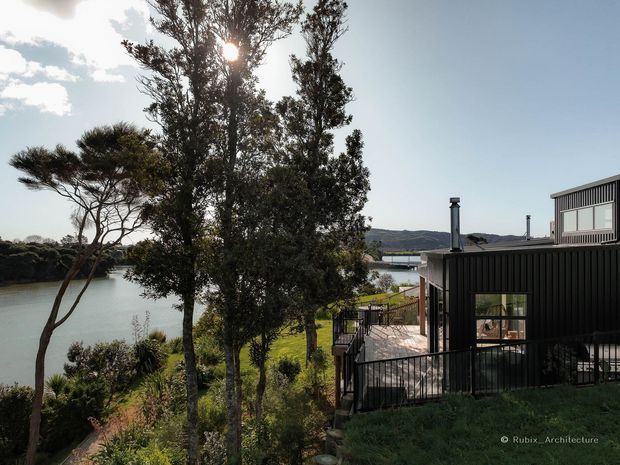 Exterior view of a contemporary dark-clad house positioned beside a calm body of water. The building features vertical ribbed metal-looking cladding, large glazed openings, a raised timber deck with black metal balustrades, and roof-mounted chimneys or flues. Mature trees and landscaped vegetation frame the waterfront setting, with hills visible across the water.