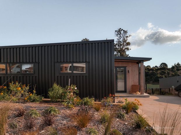 Exterior view of a compact contemporary single-storey building with dark vertically ribbed cladding, a low flat roof, and a contrasting timber-clad entrance volume. Native-style landscaped planting and a paved terrace or driveway occupy the foreground, with surrounding trees visible beyond.
