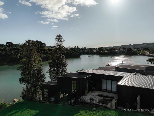 A contemporary dark-clad low-rise house positioned on a landscaped sloping site overlooking a broad body of water. The building has a strongly articulated, flat-roofed composition with corrugated metal roof sections, large glazed openings, and a terrace or courtyard area. Trees, hills, and neighbouring development are visible across the water.