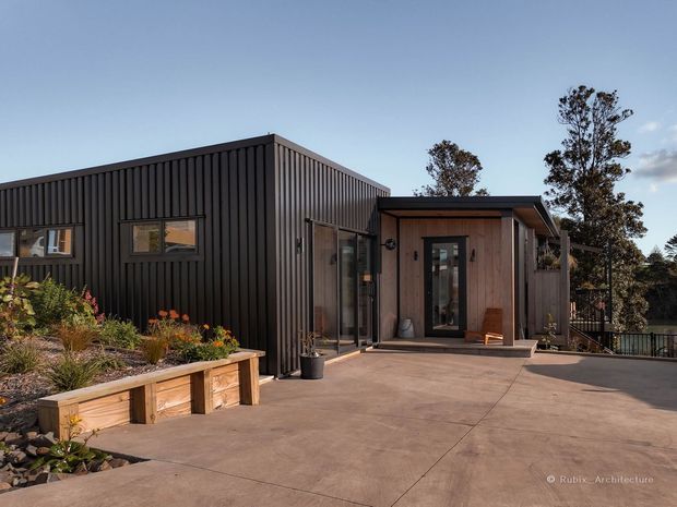 Exterior view of a contemporary single-storey building with a dark vertically ribbed facade, a timber-clad entrance volume, large glazed doors and windows, and a broad concrete terrace. Raised timber garden planters and landscaped planting occupy the foreground, with trees and a railing visible beyond.