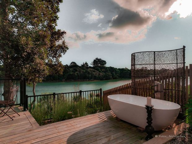 Outdoor timber deck beside a tranquil body of water, featuring a freestanding white bathtub, black decorative candle stand, metal railing, timber privacy screening, and lush vegetation. The scene has a relaxed, resort-like atmosphere with dramatic evening light and a landscaped waterfront setting.