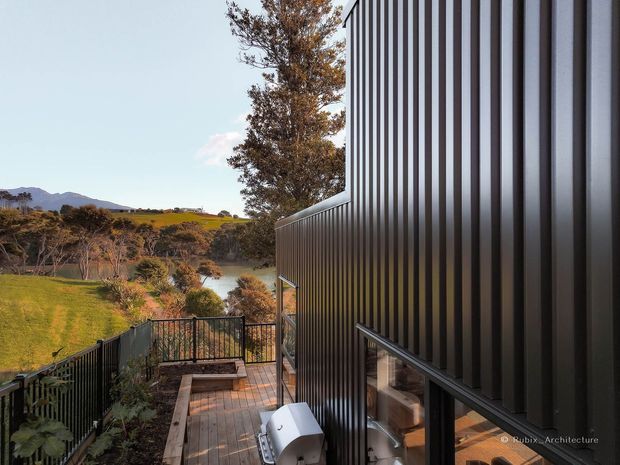 Exterior architectural photograph of a contemporary dark-clad building on a landscaped, sloping rural site. Vertical ribbed metal cladding forms the dominant facade, with large windows, a timber deck and walkway, black metal railings, and a view across green rolling farmland toward distant hills.