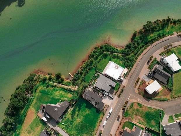 Aerial view of a residential neighbourhood beside a broad green-blue body of water. Detached homes, landscaped lots, shoreline vegetation, a small waterfront access path, and curving roads are visible from above.