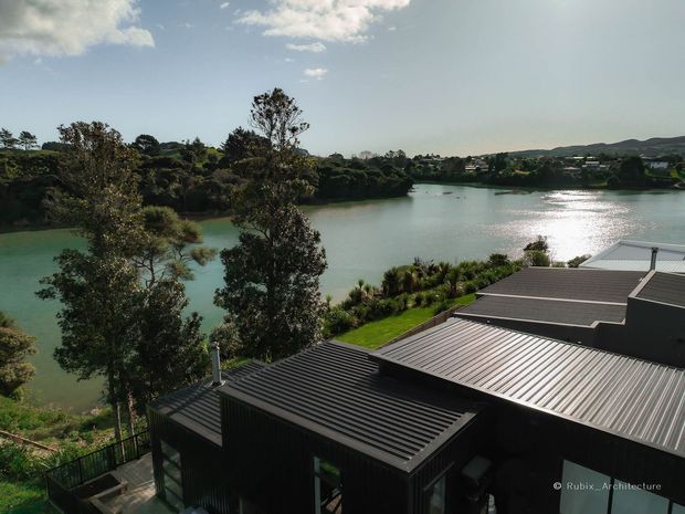 Elevated exterior view of a dark, contemporary metal-clad building with multiple corrugated metal roof planes, overlooking a calm inland waterway and landscaped residential surroundings. The image emphasizes the roofline, waterfront setting, and integration with the leafy site.