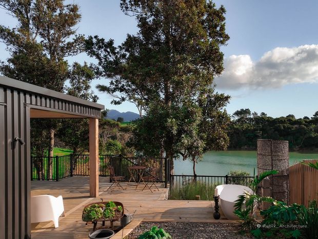 A landscaped outdoor timber deck and terrace beside a calm body of water, with a covered open-sided structure, black railings, outdoor seating, potted plants, and screening. Trees and distant hills form the backdrop, creating a relaxed waterfront entertaining area.