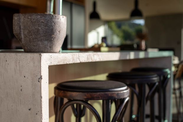 Close-up view of a contemporary kitchen or bar island with a raw concrete waterfall-style edge and benchtop. Dark metal pendant lights hang above, with several dark stools tucked beneath the overhang. A stone planter sits on the counter, and glazing in the background brings in greenery and daylight.