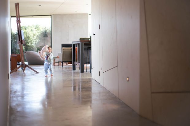 A minimalist interior hallway opening into a bright living area. The space features polished concrete flooring, smooth pale walls with flush concealed doors, recessed ceiling lighting, and large glazing overlooking greenery. A child and a few furnishings are visible in the background, with a timber easel near the window.