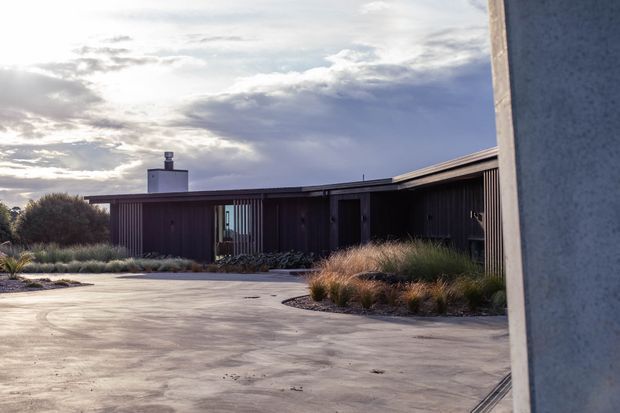 Exterior view of a low, single-storey modern residence arranged around a broad concrete-paved courtyard. The dark timber-clad building has a long, low roofline, vertical screening and glazing, and a simple chimney or rooftop vent. Native grasses and restrained planting soften the courtyard edges, while a tall pale concrete wall frames the right side. Dramatic late-day cloud and sunlight create a moody yet refined atmosphere.